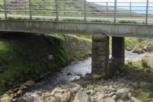 A photo of the bridge span over a small rocky river with two piers on either side of the middle of the span. It is in a rural area