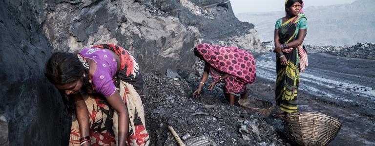 Three women work in a coal mine with a truck appearing in the distance behind them.