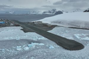 A short runway with snow covered ground to either side and snowy mountains and sea behind.