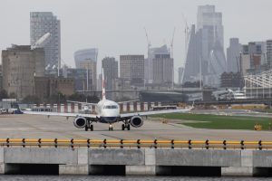 A stock photo of a passenger jet on the tarmac at London City Airport with the City of London skyscrapers in the background